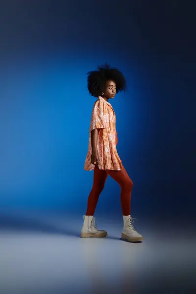 stock image stylish look of african american model in patterned shirt and orange dress posing on blue backdrop