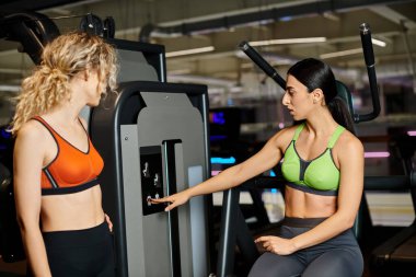 Güçlü bayan koç, spor salonunda, omuz presi makinesinin yanındaki sarışın müşterisine danışıyor.