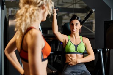 Sportif esmer kadın koç, spor salonunda, omuz presi makinesinin yanındaki sarışın müşterisine danışıyor.