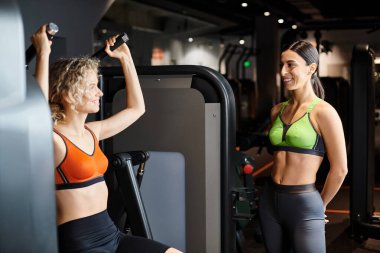 Enerjik bayan koç, spor salonunda, omuz presi makinesinin yanındaki sarışın müşterisine danışıyor.