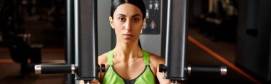 Spor giyimli güzel bir kadın göğüs pres makinesinde egzersiz yapıyor ve kameraya bakıyor.