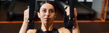 Spor kıyafetleri içinde esmer, enerjik bir kadın spor salonunda barfiks ekipmanı ve pankart kullanıyor.