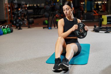 Spor elbiseli çekici esmer kadın spor salonundayken spor minderinde ağırlık tabağıyla oturuyor.
