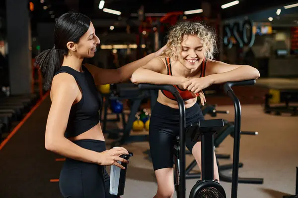 Stock image jolly athletic female coach helping her beautiful client to use cross trainer in gym, water bottle