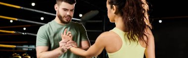 Bir erkek antrenör, spor salonunda bir kadına kendini savunma teknikleri gösterir..