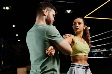 Bir erkek antrenör, spor salonunda bir kadına kendini savunma teknikleri gösterir..
