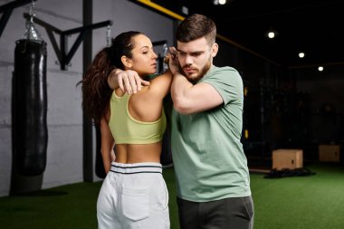 Bir erkek antrenör, spor salonunda bir kadına savunma teknikleri öğretir, dayanıklılık ve birlik gösterir..