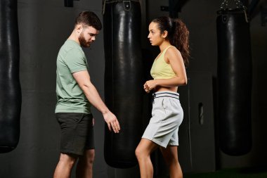 Erkek bir antrenör spor salonunda bir kadına savunma teknikleri öğretir. Takım çalışması ve güç gösterisi yapar..