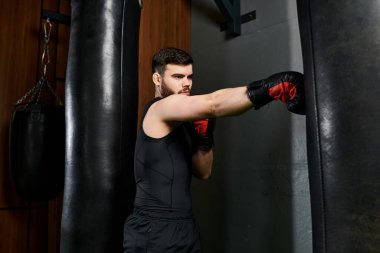 A handsome man in a black shirt delivers punches to a punching bag with red boxing gloves in a gym. clipart