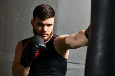 Siyah gömlekli ve boks eldivenli sakallı bir adam spor salonundaki kum torbasına yumruk atıyor..