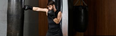 Siyah gömlekli, siyah eldivenli, sakallı yakışıklı bir adam ringde kum torbasıyla boks yapıyor..