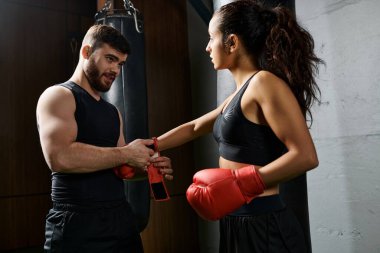 Erkek bir antrenör spor salonunda boks eldiveni giyer ve antrenman yaparken aktif giyinen esmer bir sporcunun yanında durur..