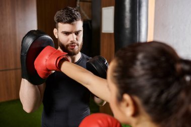 Erkek bir antrenör, spor salonunda boks yaparken etkin giyinmiş esmer bir sporcuya yardım eder..