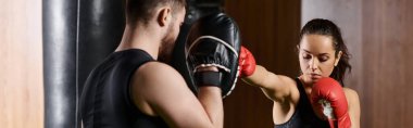 Spor salonunda boks yapan esmer ve sporcu bir kadının yanında duran bir erkek antrenör..
