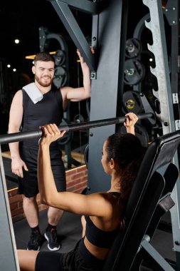 Erkek bir antrenör, esmer bir sporcuya spor salonunda çömelme hareketleri yaparken rehberlik eder..