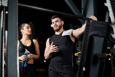 Spor salonunda birlikte duran erkek bir spor eğitmeni ve esmer bir kadın..