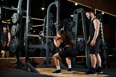 Kişisel antrenör ve esmer bir sporcu eş zamanlı olarak iyi donanımlı bir spor salonunda çömelme hareketleri yapıyor..