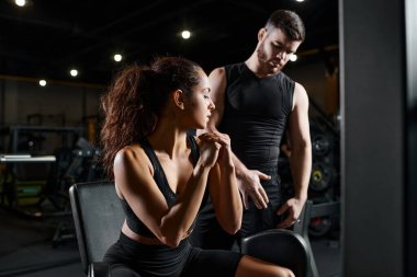 A personal trainer coaches a brunette sportswoman as they stand side by side in a gym, focusing on fitness and strength training. clipart