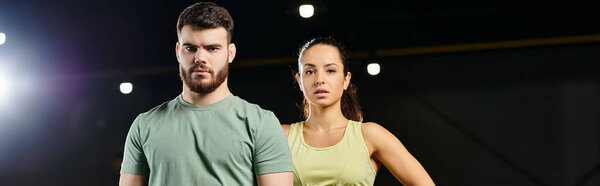 A male trainer and woman in a gym, both standing confidently next to each other.