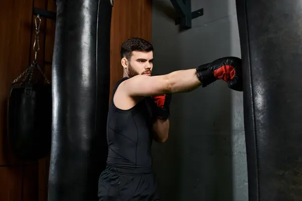 A handsome man in a black shirt delivers punches to a punching bag with red boxing gloves in a gym.