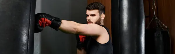A bearded man in a black shirt and red boxing gloves fiercely punches a bag in a gym.