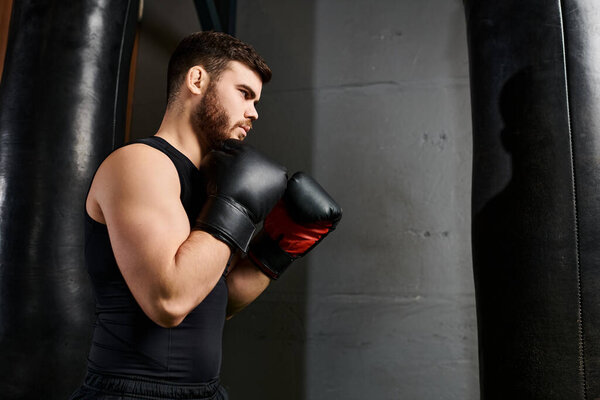 Красивый мужчина с бородой в черной майке бьет боксерскую сумку в спортзале, в ярких красных перчатках..