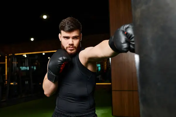stock image A bearded man in a black shirt trains in a gym, throwing punches at a heavy bag with black boxing gloves.