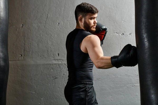 A handsome man with a beard wearing a black shirt and red boxing gloves, punching a bag in a gym.