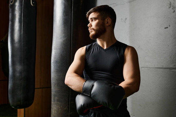 Бородач в черной майке и боксерских перчатках бьет сумку в спортзале..