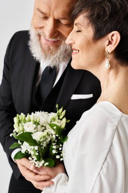 Smokinli orta yaşlı damat, özel günlerini kutlamak için bir stüdyo ortamında gelinliğinin yanında buket tutuyor..