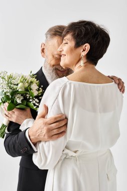 Middle-aged bride and groom in wedding attire hugging tightly while holding flowers, celebrating their special day in a studio setting. clipart