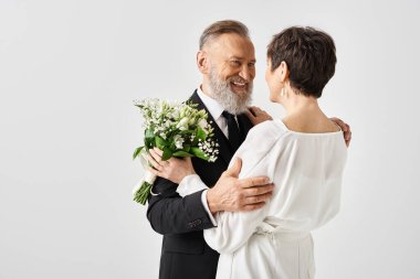 Smokinli orta yaşlı bir damat beyaz elbiseli gelinini nazikçe kucaklıyor ve stüdyo ortamında özel günlerini kutluyor..