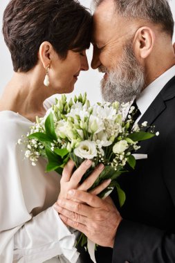 Düğün kıyafetleri içinde orta yaşlı gelin ve damat ellerinde güzel bir buket çiçekle stüdyo ortamında özel günlerini kutluyorlar..