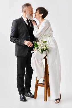 A middle-aged bride and groom, dressed in wedding gowns, sit together on a chair, celebrating their special day in a studio setting. clipart