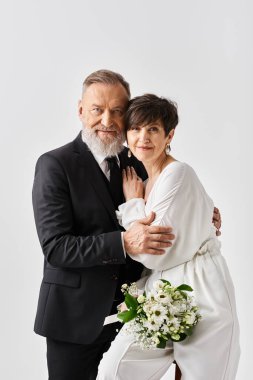 Beyaz elbiseli orta yaşlı bir gelin ve takım elbiseli bir damat stüdyodaki özel günlerinde şefkatle kucaklaşırlar..
