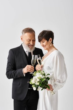 Middle-aged bride and groom elegantly stand side by side, holding champagne glasses in a studio setting, celebrating their special day. clipart