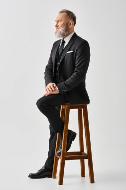 Orta yaşlı damat şık bir takım elbise içinde stüdyo ortamında bir taburede oturuyor ve düğün gününde sonsuz bir cazibe yayıyor..