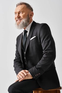 A middle-aged groom in an elegant suit sits on a stool in a studio setting on his wedding day. clipart