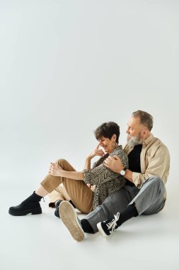 Orta yaşlı, şık giyinmiş bir çift stüdyo ortamında yan yana oturmuş, sofistike ve birlik içinde..