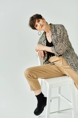 Kısa saçlı şık, orta yaşlı bir kadın stüdyo ortamında beyaz bir taburede otururken zarafet saçıyor..