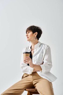 Middle-aged woman in stylish attire sits on stool holding a cup of coffee in a serene studio setting. clipart