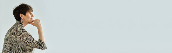 Женщина средних лет, стильная в одежде, стоит с рукой на подбородке, потерявшись в мыслях в студии.