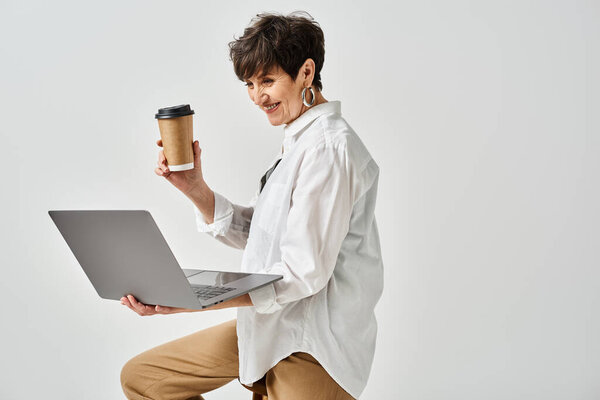 Женщина средних лет в стильной одежде, держащая чашку кофе и работая над ноутбуком в студии.