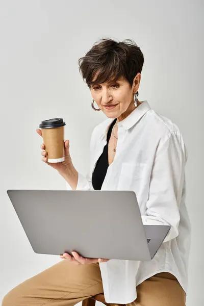 Middle aged woman multitasking, holding a coffee cup and laptop in a stylish studio setting.