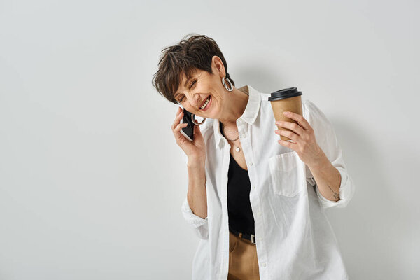 A middle-aged woman with short hair multitasking, holding coffee cup and chatting on cellphone in a trendy studio.