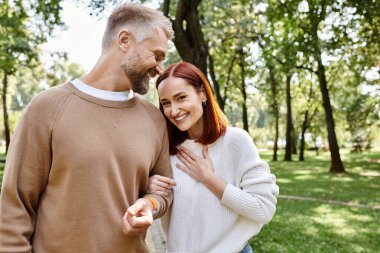 Bir adam ve bir kadın günlük giysiler içinde huzurlu bir parkta yan yana duruyorlar..