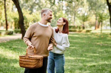 Bir çift, gündelik giyinmiş, yeşil alanlarla çevrili bir parkta rahatça yürüyor..