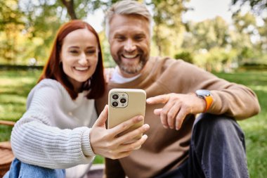 Bir adam ve bir kadın günlük giysiler içinde parkta yürürken selfie çekiyorlar..