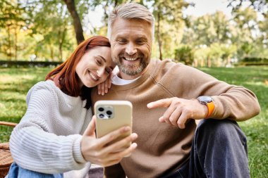 Sıradan giyinmiş bir çift romantik bir şekilde parkta cep telefonuyla selfie çekiyor..