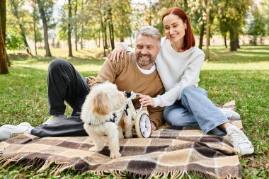 Bir erkek ve bir kadın, huzurlu bir parkta bir köpekle battaniyenin üzerinde rahatlar..